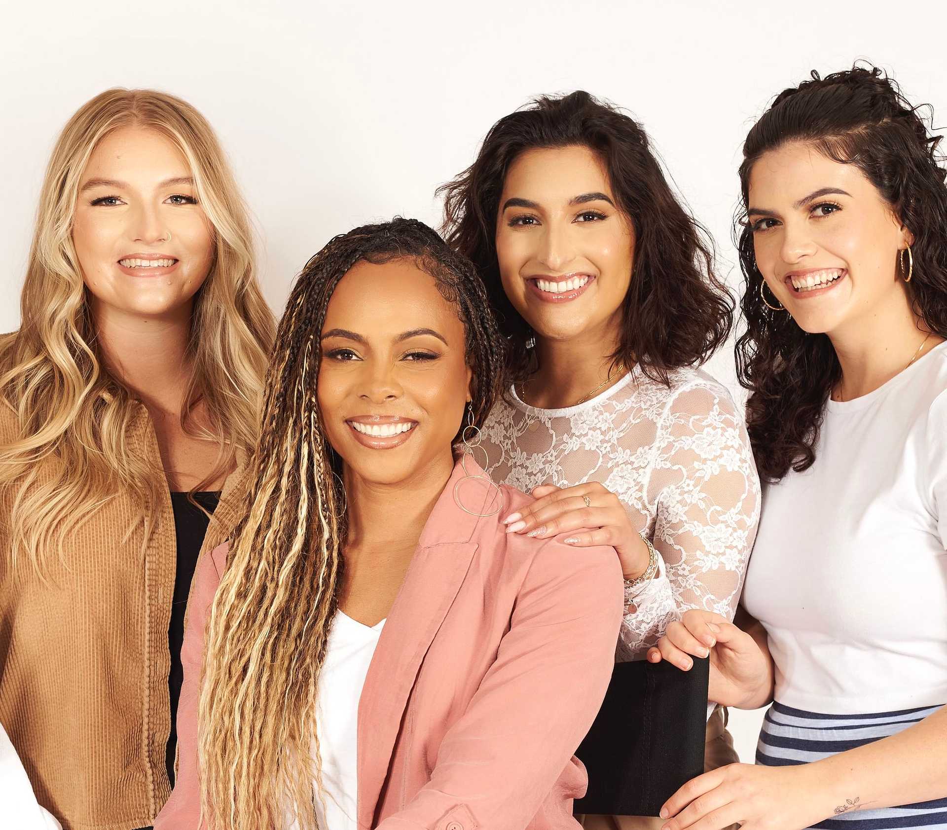 Four women smiling together against a white background, showing diverse hairstyles and outfits.
